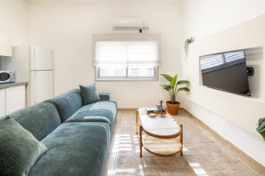 A modern living room with a sofa, a coffee table, a TV, and a window with blinds. - 3BR + 3 Baths near Gordon Beach by SeaNRent (Tel Aviv)