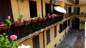 Un patio estrecho con un edificio amarillo, plantas en macetas y un balcón con sombrilla.