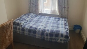 Una cama pequeña y bien hecha con colcha azul y blanca a cuadros, una silla de mimbre y una ventana con cortinas florales.
