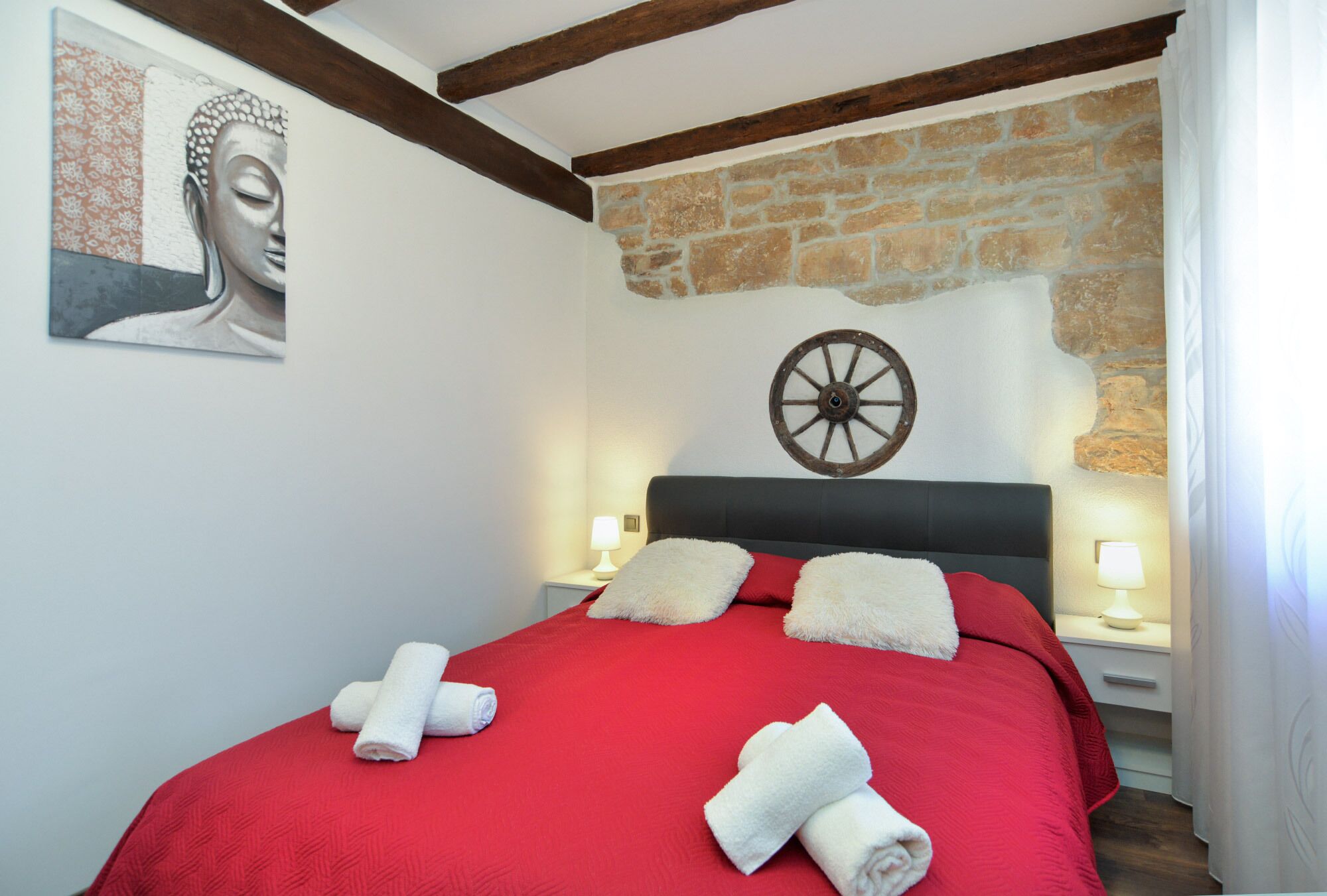 Una cama bien tendida con ropa de cama roja y toallas blancas, un cabecero de madera, una pared de piedra y una rueda fijada a la pared.