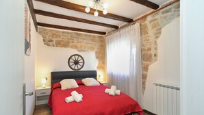 A bedroom with a stone wall, wooden beams, a bed with a red bedspread, two white pillows, and a wall-mounted wheel.