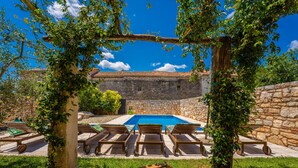 Un edificio de piedra con piscina, rodeado de sillones de madera y follaje.