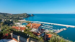 Una marina costiera con imbarcazioni ormeggiate, un molo che si protende nell'acqua e edifici con tetti in tegole rosse.