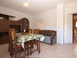 Comedor con una mesa dispuesta para cuatro personas, rodeada de sillas de madera. Un mantel con estampado floral cubre la mesa, y se observa un aparador con puertas de cristal al fondo.