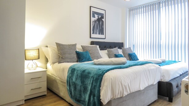 A modern bedroom with a large bed, bedside tables, a lamp, and a window with blinds. - Tudors eSuites Birmingham Canalside with Gated Parking (Birmingham)