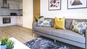 A living room with a grey sofa, a blue rug, and a coffee table. There are two framed artworks on the wall and a potted plant on the table. - Tudors eSuites Birmingham Canalside with Gated Parking (Birmingham)