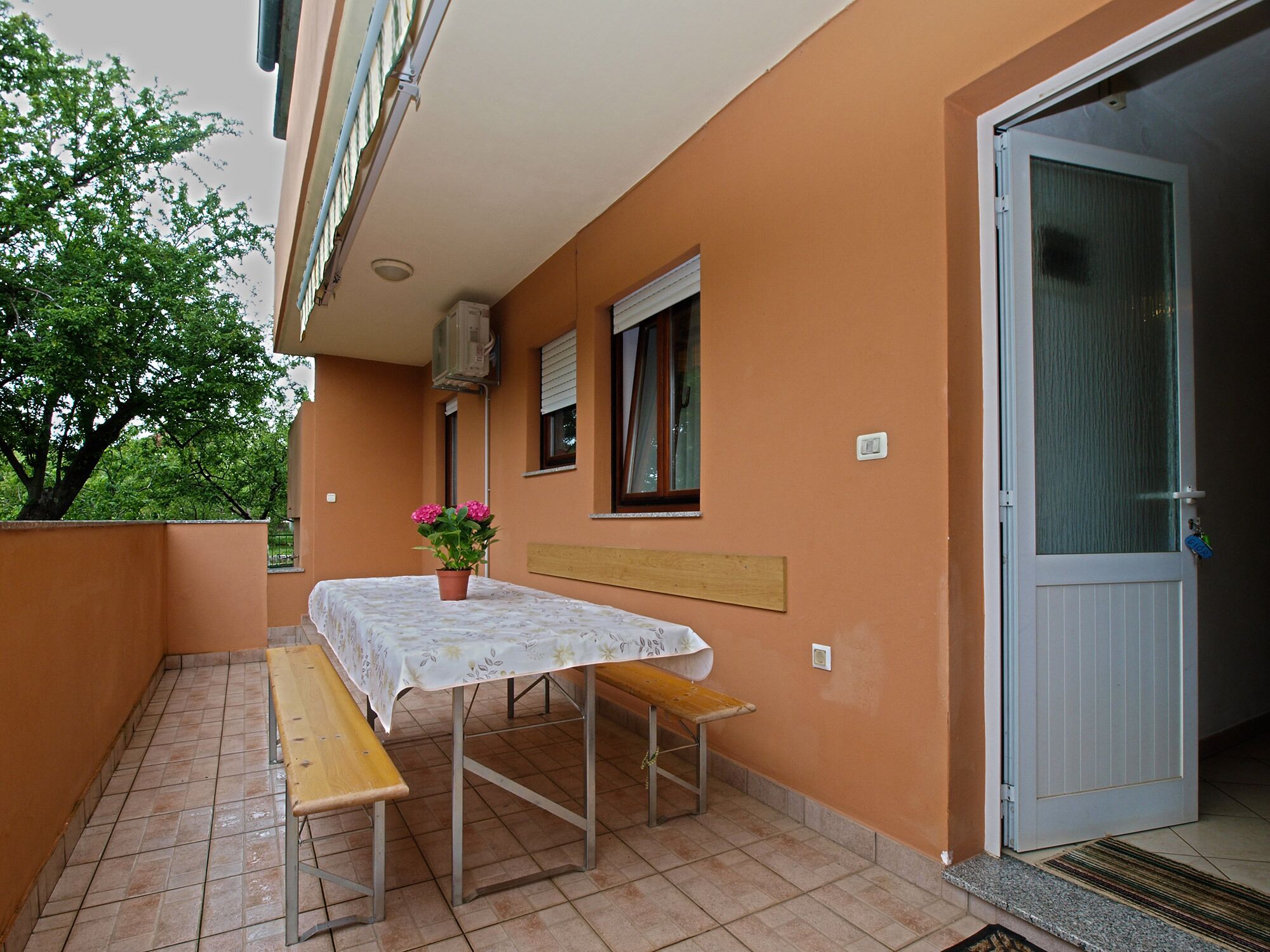 A balcony with a table and benches, a potted plant, a door, and a window with blinds.