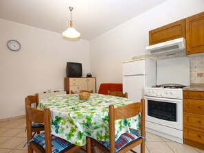 Un comedor con una mesa cubierta con mantel floreado, rodeada por sillas de madera con cojines azules y rojos. Un televisor está montado en un mueble de madera sobre la mesa.
