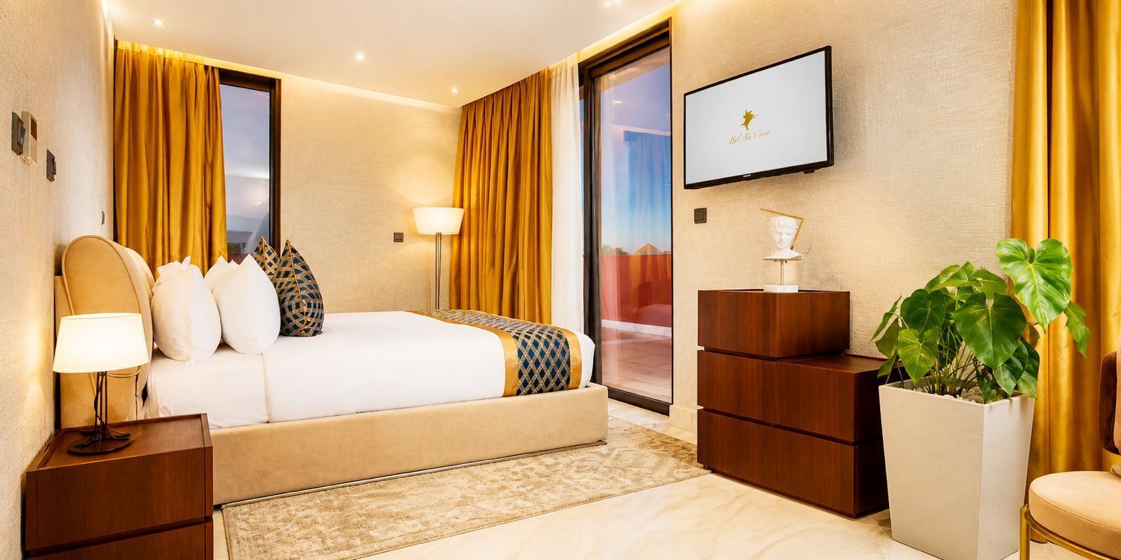 A hotel room with a bed, bedside tables, a TV, a desk, and a large plant.