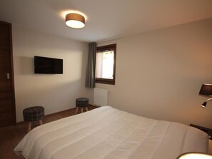 Quarto com cama, televisão fixada na parede, janela com cortinas e abajur.