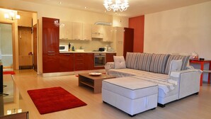 Een moderne woonkamer met een bank, een poef en een rood vloerkleed. Er is een keukenhoek met kasten en een magnetron.