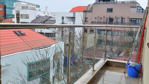 Een balkon met een metalen hekwerk, een blauwe emmer en een rood dak in de verte.