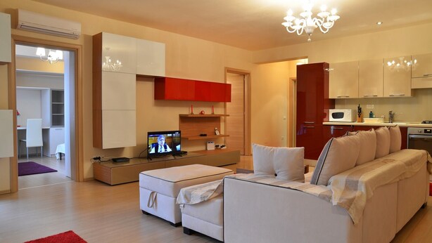 Een moderne woonkamer met een bank, een poef en een televisie waarop een nieuwsuitzending wordt vertoond.