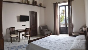 A hotel room with a bed, two chairs, a desk with a TV, a window with curtains, and a view of palm trees. - Agorà Panoramic Rooms (Sciacca)