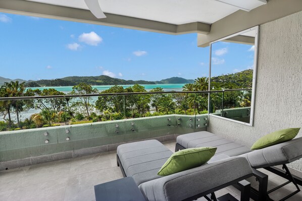 A balcony with a view of a turquoise sea and green hills. There are two grey lounge chairs with green cushions.
