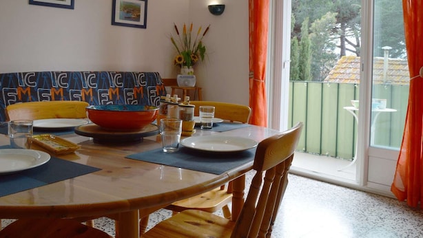 A dining area with a wooden table, chairs, and a large bowl. There are two framed pictures on the wall, a vase with sunflowers, and a window with orange curtains. - Cozy Apartment with Parking, 100m from Beach - Argelès-sur-Mer (Argelès-sur-Mer)