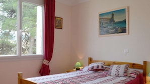 A bedroom with a wooden bed, a painting of a lighthouse, a window with red curtains, and a small bedside table with a lamp. - Cozy Apartment with Parking, 100m from Beach - Argelès-sur-Mer (Argelès-sur-Mer)