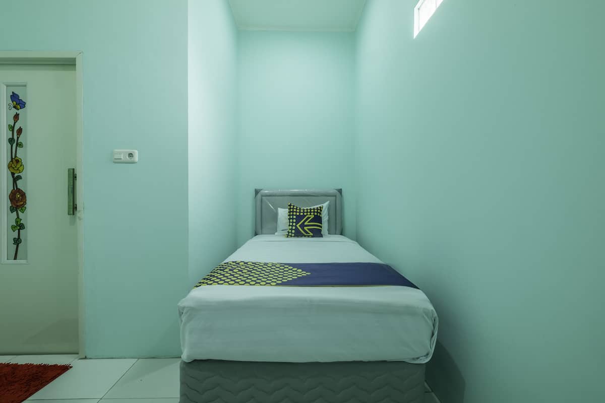 A narrow room with a bed, a door with a floral decoration, and a thermostat on the wall.