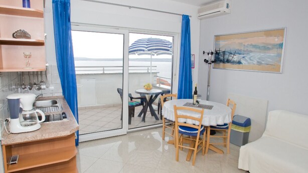 A room with a kitchenette, a dining area with a table and chairs, a white sofa, and a view of the sea through a sliding glass door. - Apartments Spommy (Crikvenica)