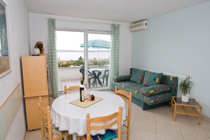 A room with a round table, wooden chairs, a sofa with colorful cushions, a small side table, and a potted plant.