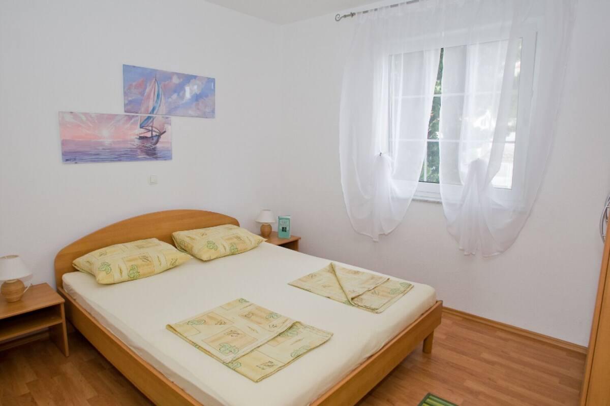 Quarto com cama de madeira, duas almofadas, mesa de cabeceira com abajur e janela com cortinas leves.
