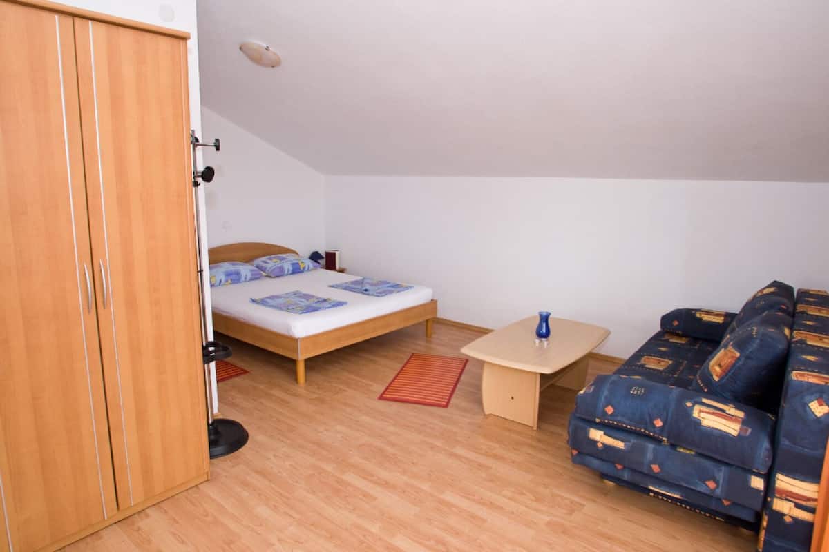 Quarto pequeno e bem iluminado com um guarda-roupa de madeira, uma cama de solteiro com cabeceira de madeira, uma mesinha e um sofá azul.