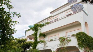 A white building with multiple balconies, surrounded by lush greenery and a stone wall. - Apartments Spommy (Crikvenica)