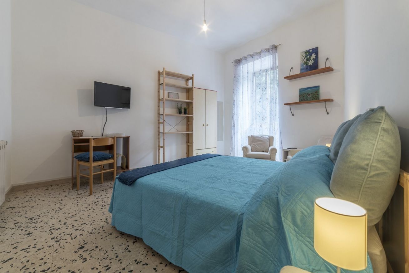 A bedroom with a bed, a desk, a chair, a TV, shelves, and a window.