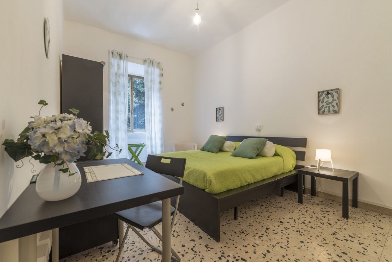A bedroom with a bed, a desk, a chair, a bedside table, a lamp, and a window with curtains.