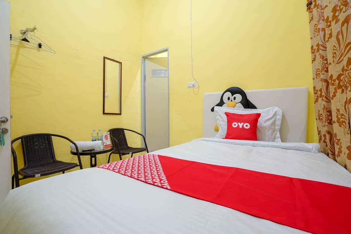 Une chambre d’hôtel avec un lit, une petite table sur laquelle sont posées des bouteilles, deux chaises, un étendoir et une porte.