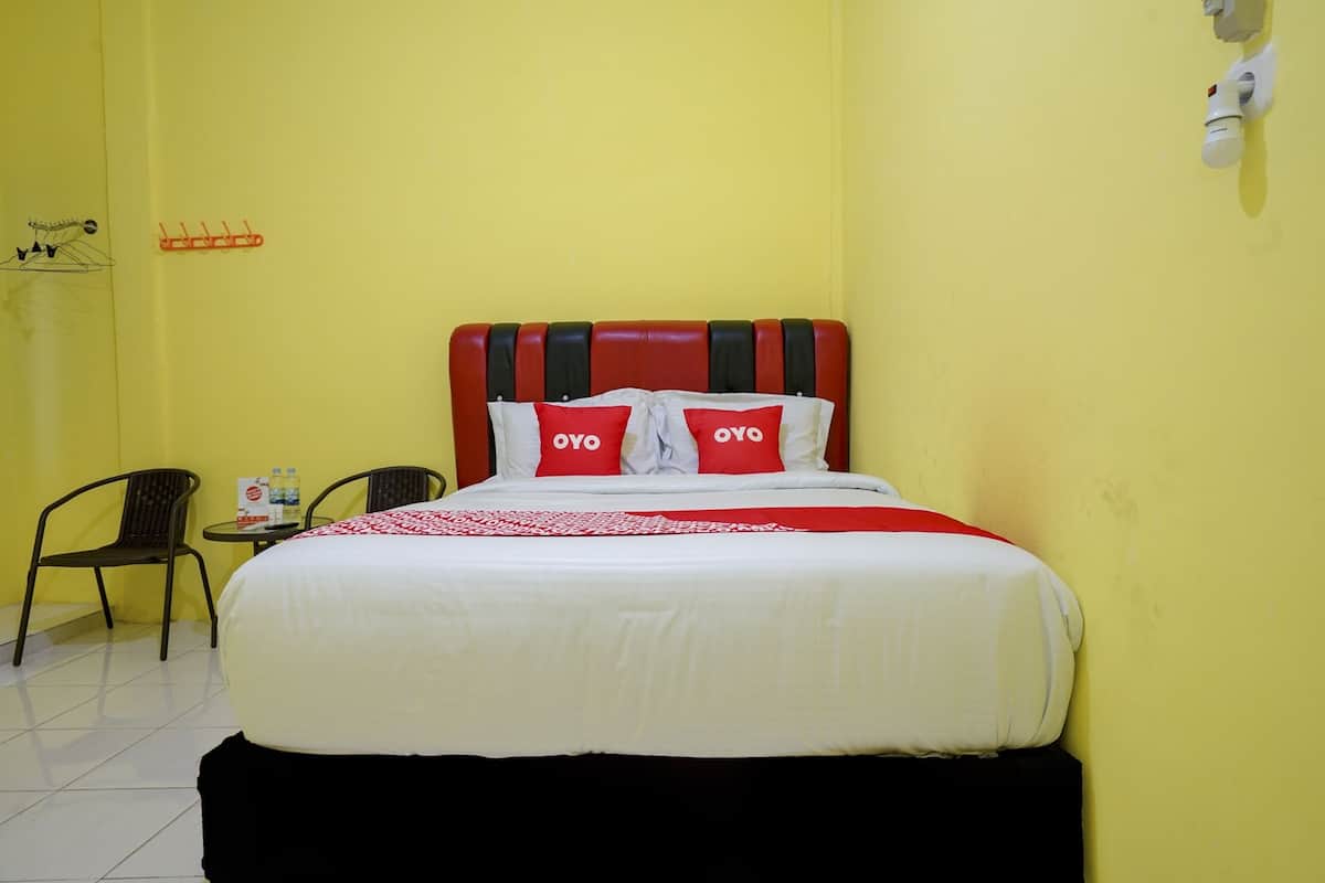 Une chambre d’hôtel avec un lit, deux chaises, une petite table sur laquelle se trouvent une bouteille et une boîte, et un luminaire fixé au mur.