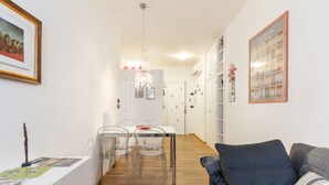 Una sala moderna con un sofá gris, un comedor con una mesa blanca y sillas transparentes, y un pasillo que lleva a una cocina.
