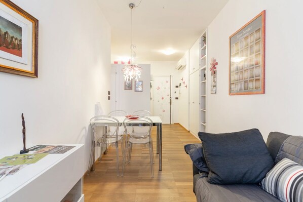 A modern living room with a gray sofa, a dining area with a white table and clear chairs, and a hallway leading to a kitchen.
