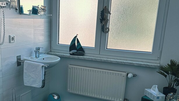 A modern bathroom with a toilet, sink, and a small sailboat decoration on the windowsill. - ORCA Hotel am Elbschloss (Dresden)