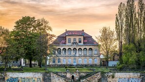 A historic building with arched windows and a red roof, situated by a river with a bridge leading to it. - ORCA Hotel am Elbschloss (Dresden)