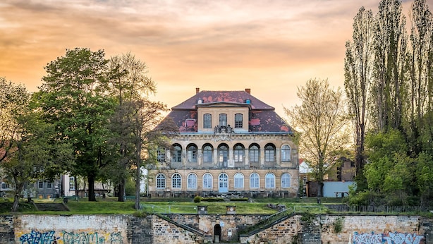 A historic building with arched windows and a red roof, situated by a river with a bridge leading to it. - ORCA Hotel am Elbschloss (Dresden)