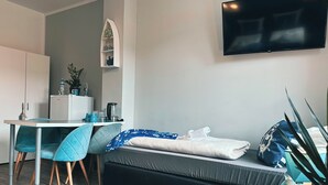 A modern room with a dining table, chairs, a sofa, a TV, and a small kitchen area. - ORCA Hotel am Elbschloss (Dresden)