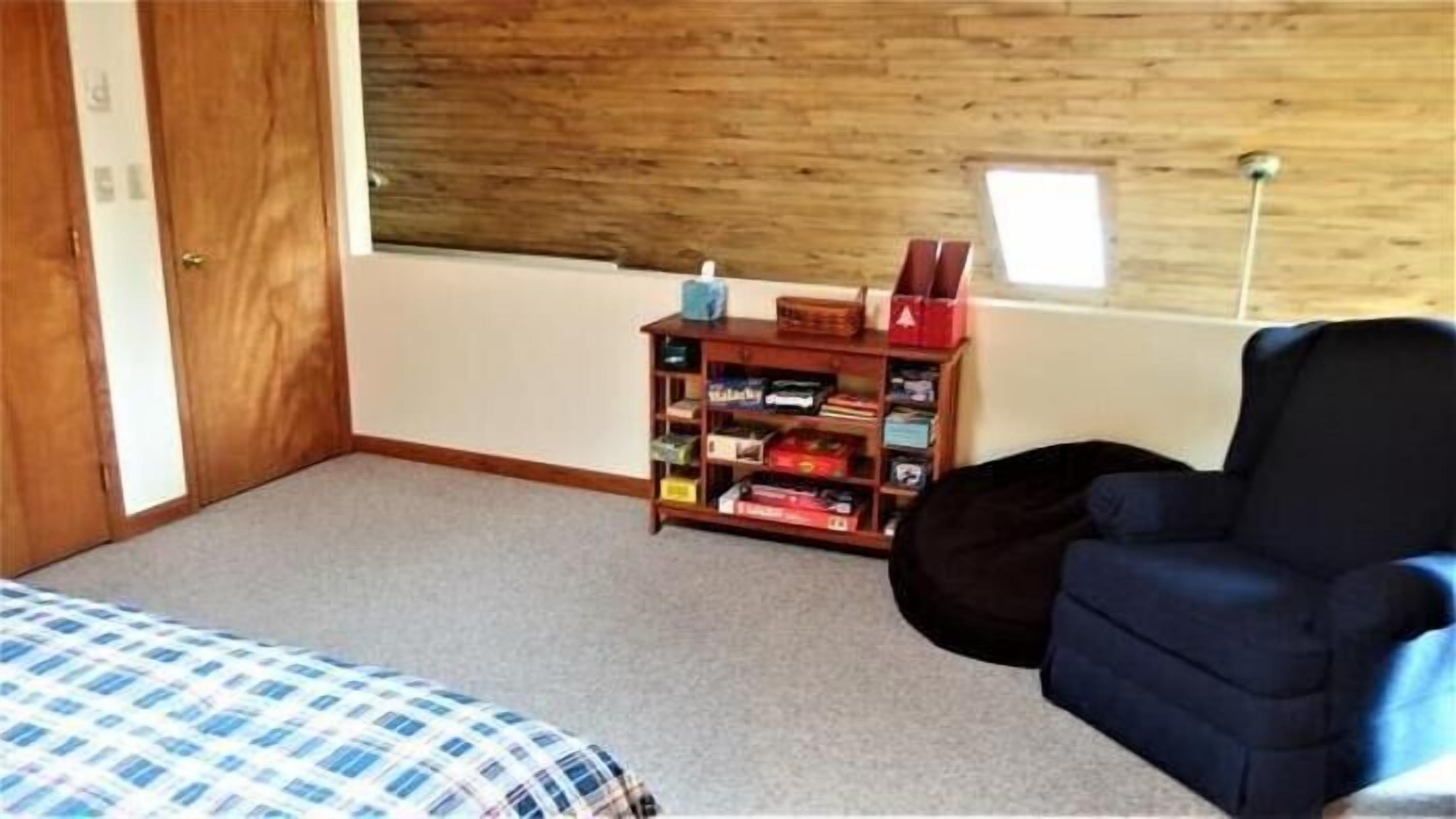 A bedroom with a wooden headboard, a bookshelf, a blue armchair, and a bed with a checkered bedspread.