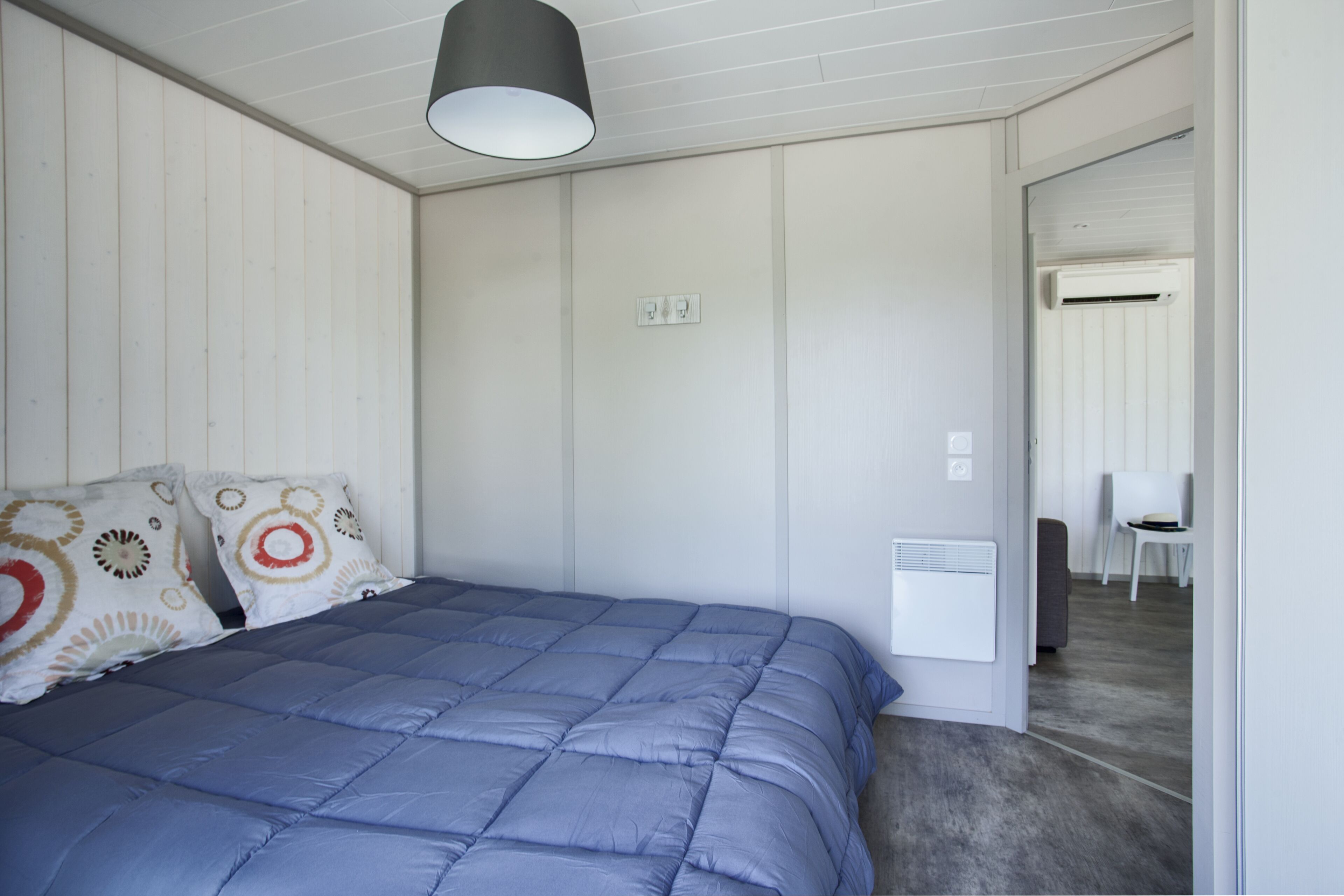 A bedroom with a blue bedspread, white walls, a ceiling light, and a white air conditioning unit.