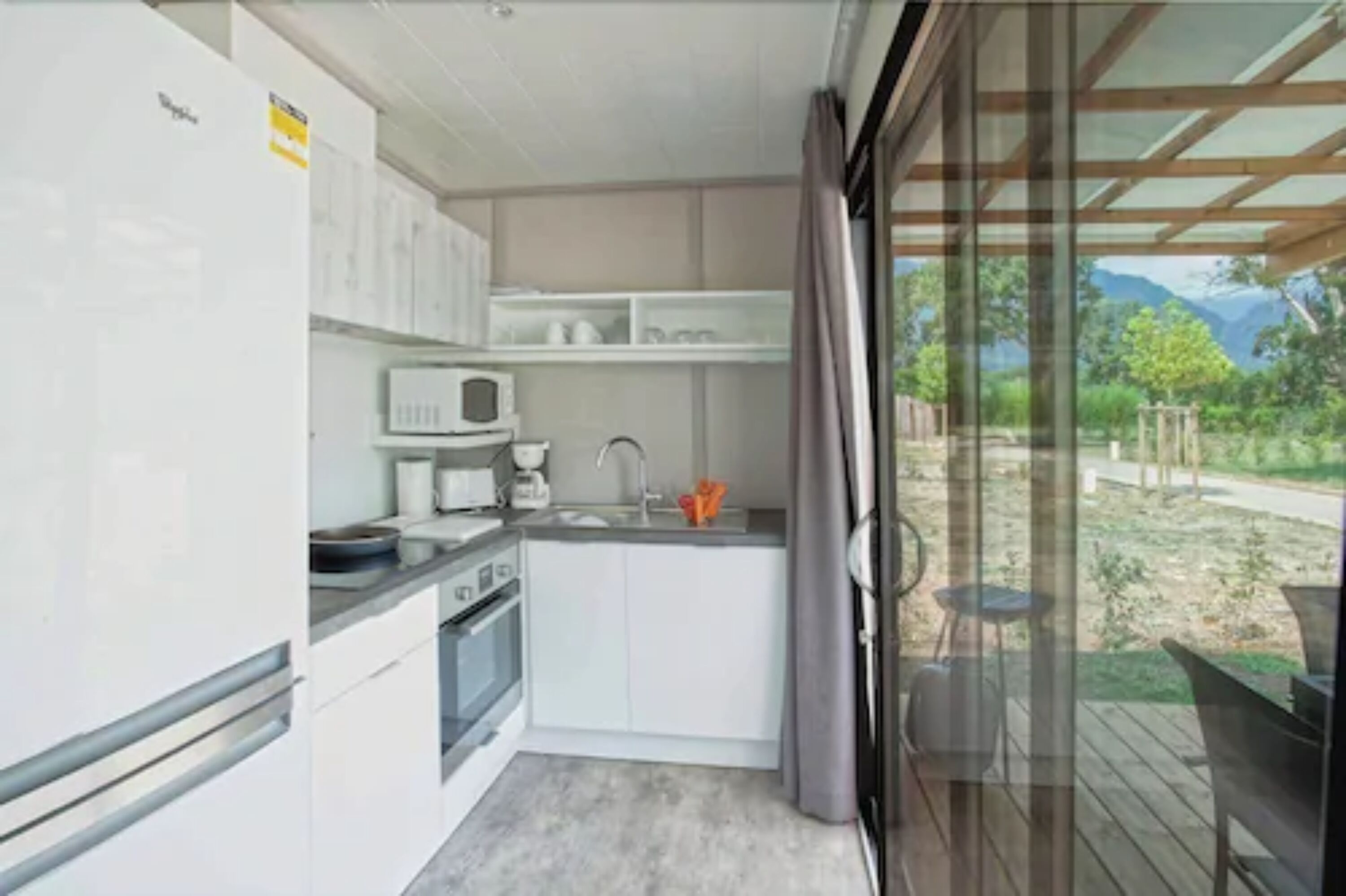 A compact kitchen with white cabinets, a microwave, and a sink. It has a sliding glass door leading to an outdoor area with a view of greenery and mountains.
