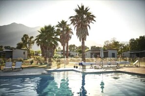 Una piscina con sillas de descanso y palmeras, rodeada de edificios y una montaña al fondo.