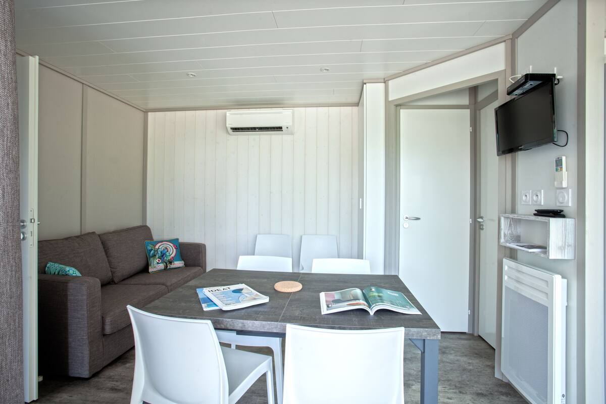 Una habitación con una mesa gris, sillas blancas, un sofá marrón con un cojín colorido, una revista sobre la mesa y una puerta blanca con un televisor y termostato empotrados.