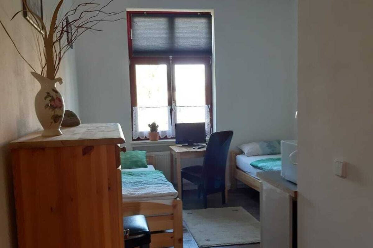 A small, narrow room with a bed, a desk, and a chair. There is a window with blinds, a potted plant, and a wooden cabinet.
