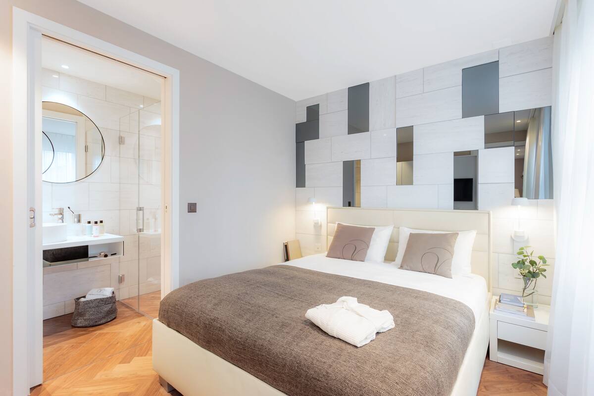 A modern hotel room with a large bed, a bedside table with a lamp, a mirror, and a bathroom with a sink and toilet.