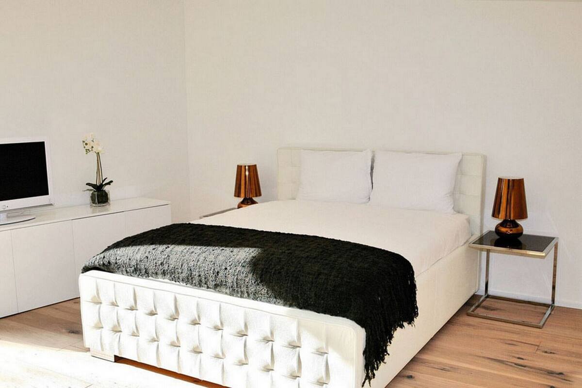 A modern bedroom with a white bed, a TV on a white cabinet, and two lamps on a side table.