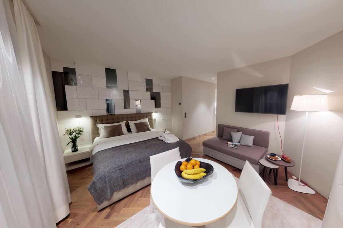 A modern hotel room with a bed, a sofa, a small table with fruit, a TV, and a lamp.