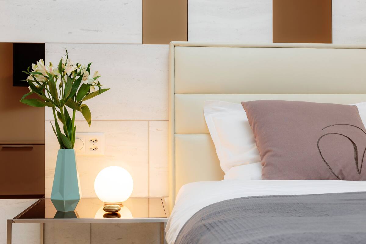 A neatly made bed with a headboard, a bedside table with a vase of flowers, and a wall-mounted light.