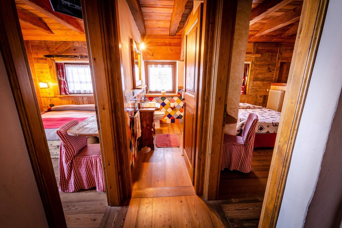Interior de una cabaña de madera con un estrecho pasillo que lleva a un dormitorio con cama y ventana.