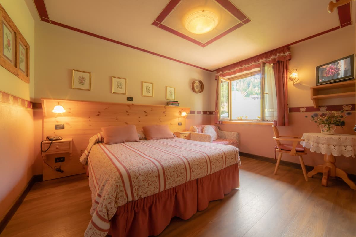 Un dormitorio con cabecera de madera, cama con cubrecama floral, mesita de noche con lámpara, una silla y una ventana con cortinas.
