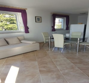 Uma sala de estar moderna com um sofá bege, uma mesa de jantar de vidro com quatro cadeiras e uma geladeira.